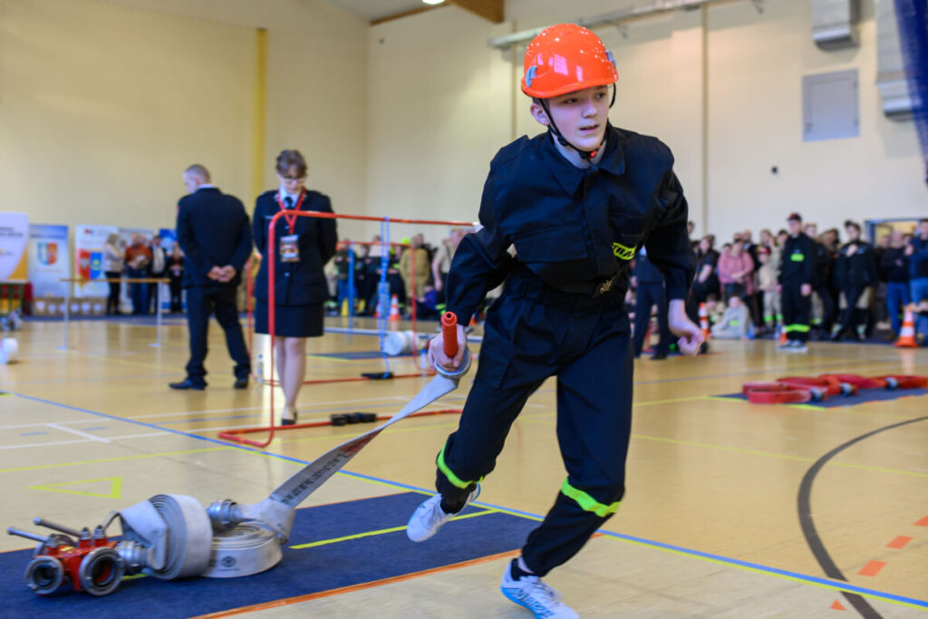 Młody uczestnik w kasku i uniformie biegnie z wężem strażackim na zawodach w hali sportowej. Tło to zebrana publiczność i sędziowie. Kolory dominujące to czarny, pomarańczowy i beżowy.