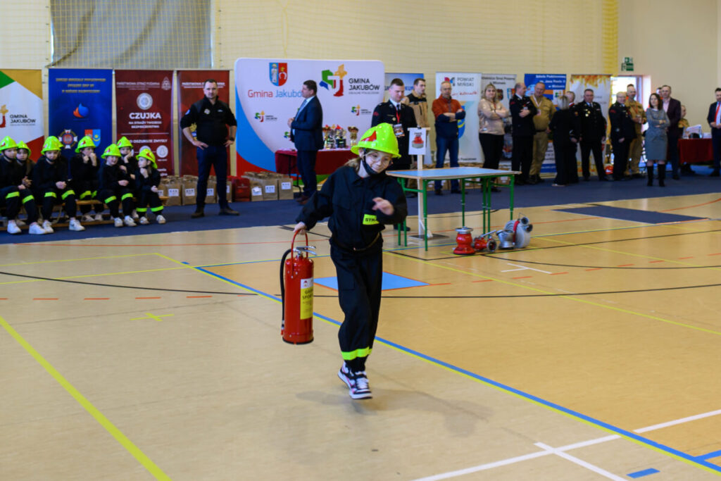 Na hali sportowej dziecko w zielonym hełmie biegnie z gaśnicą. W tle widoczni strażacy i kolorowe plakaty. Na podłodze są oznaczenia.