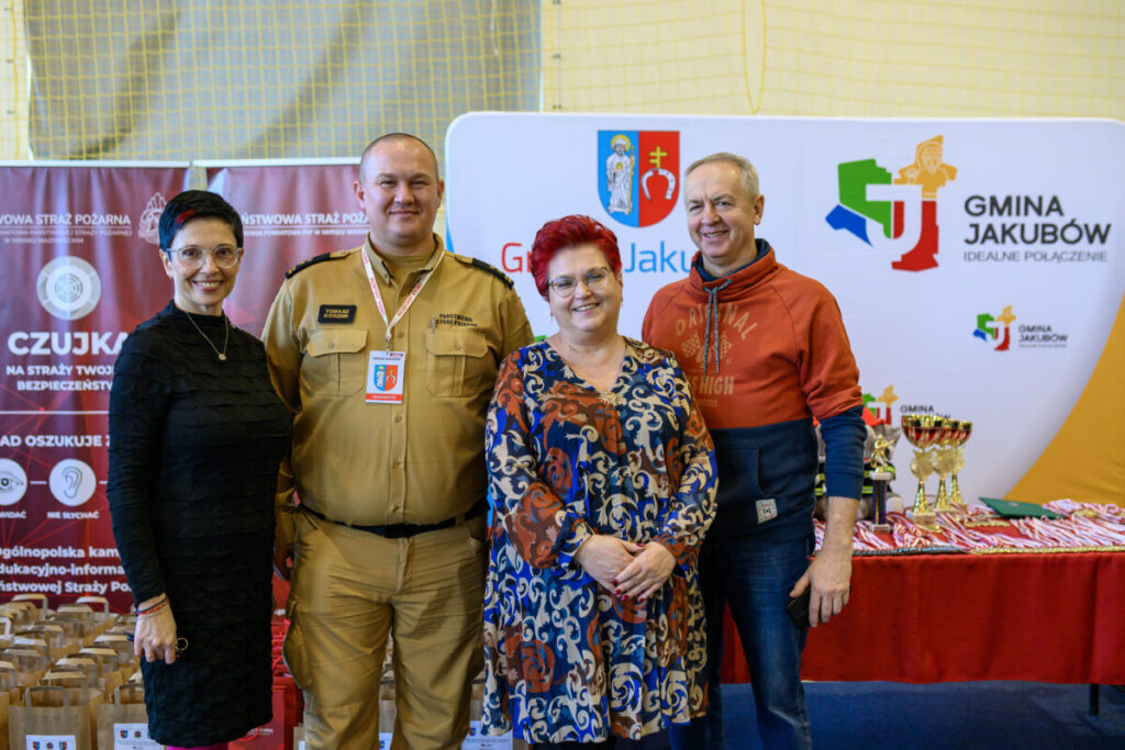 Cztery osoby pozują razem na tle banerów z logo gminy Jakubów. Jedna z osób jest ubrana w strój strażacki. Obok stoją puchary i medale na stole.