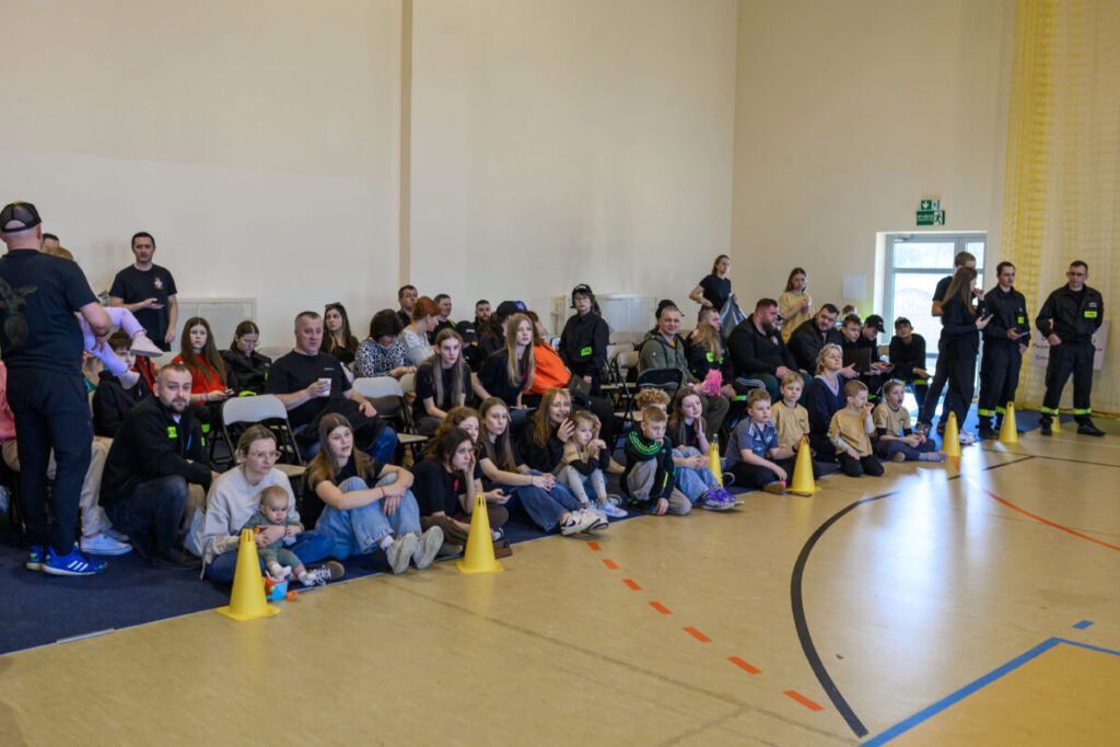 Grupa ludzi, w tym dzieci, siedzi na podłodze i krzesłach w jasnej sali gimnastycznej. Żółte pachołki stoją na drewnianej podłodze. Tło ma beżowe ściany z drzwiami przy wyjściu ewakuacy