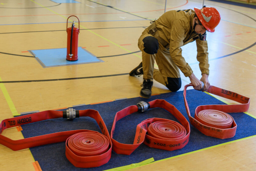Strażak w czerwonym kasku układa czerwony wąż strażacki na niebieskiej macie. Tło jasne, linie oznaczają strefy. Obok czerwona gaśnica.