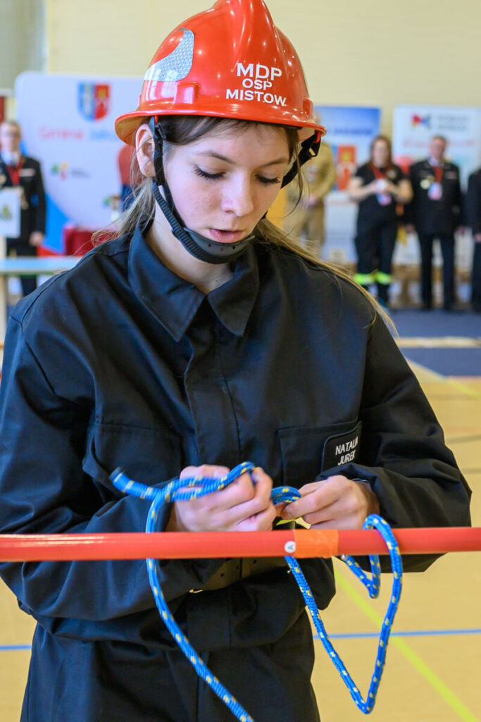 Młoda kobieta w czerwonym kasku i czarnym stroju wiąże niebieską linę. Tło jest rozmyte z postaciami. Scena odbywa się w jasnym pomieszczeniu.