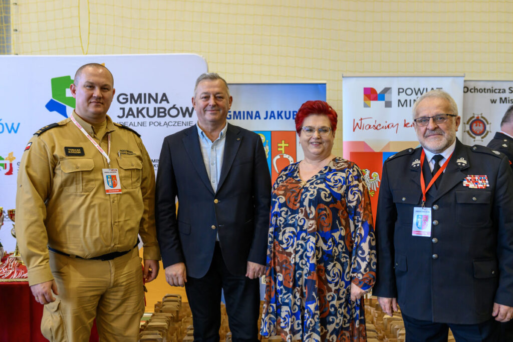 Cztery osoby na tle banerów w jasnym, zadaszonym pomieszczeniu. Noszą formalne i służbowe ubrania. Kolory ubrań to beżowy, granatowy i wzorzysty.