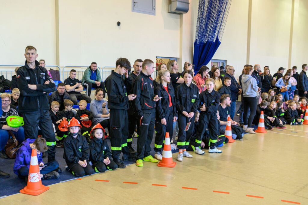 Grupa młodych ludzi w czarnych uniformach stoi w sali gimnastycznej z pomarańczowymi pachołkami. W tle siedzą rodzice i dzieci, ściana jest jasna. Obok niektórzy trzymają kaski strażackie.