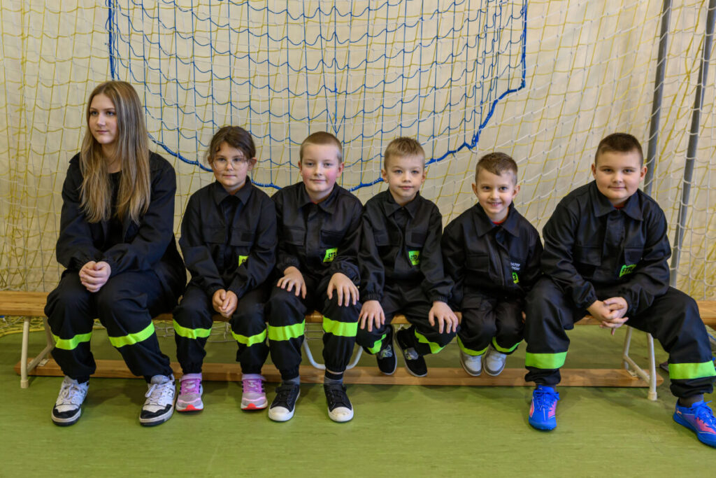 Na zdjęciu jest sześć osób w czarnych strojach z żółtymi odblaskami. Siedzą na ławce w sali gimnastycznej z siatką w tle. Podłoga jest zielona, mają kolorowe buty.