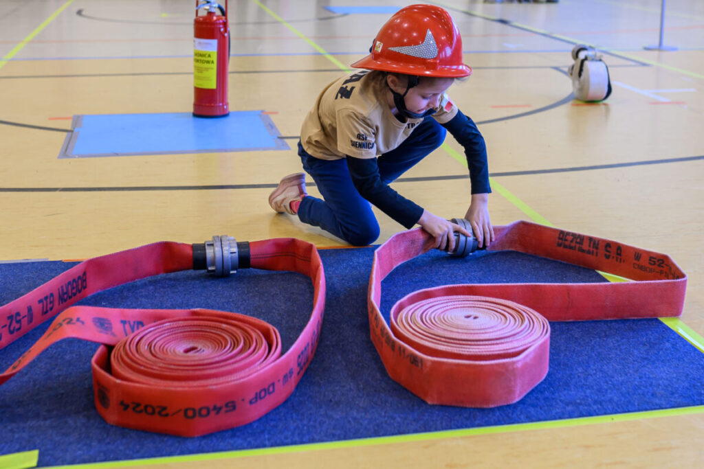 Małe dziecko w czerwonym kasku bawi się w strażaka, rozwijając czerwony wąż na niebieskiej macie na sali gimnastycznej. W tle widoczny jest czerwony gaśnica i jasne podłoże z wyznaczonymi liniami. Dziecko jest u