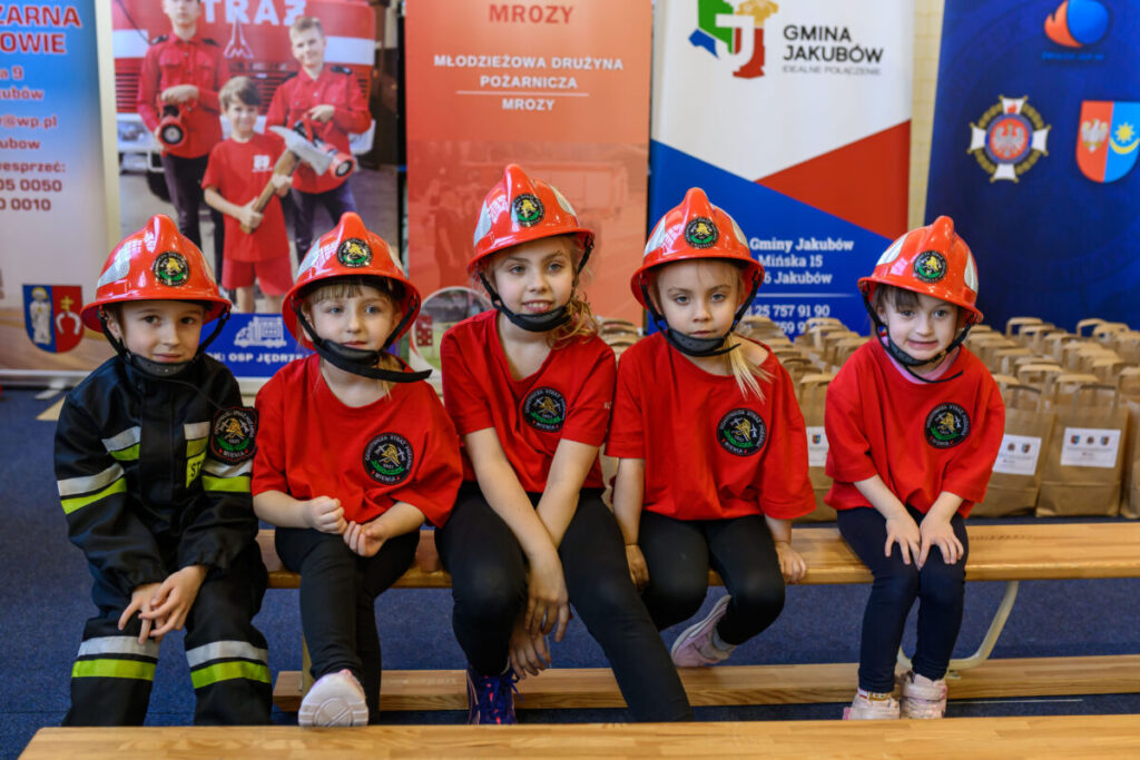 Pięcioro dzieci w czerwonych kaskach i strojach strażackich siedzi na ławce. Tło z banerami w kolorach czerwonym, niebieskim i białym. Obok nich rząd papierowych toreb.