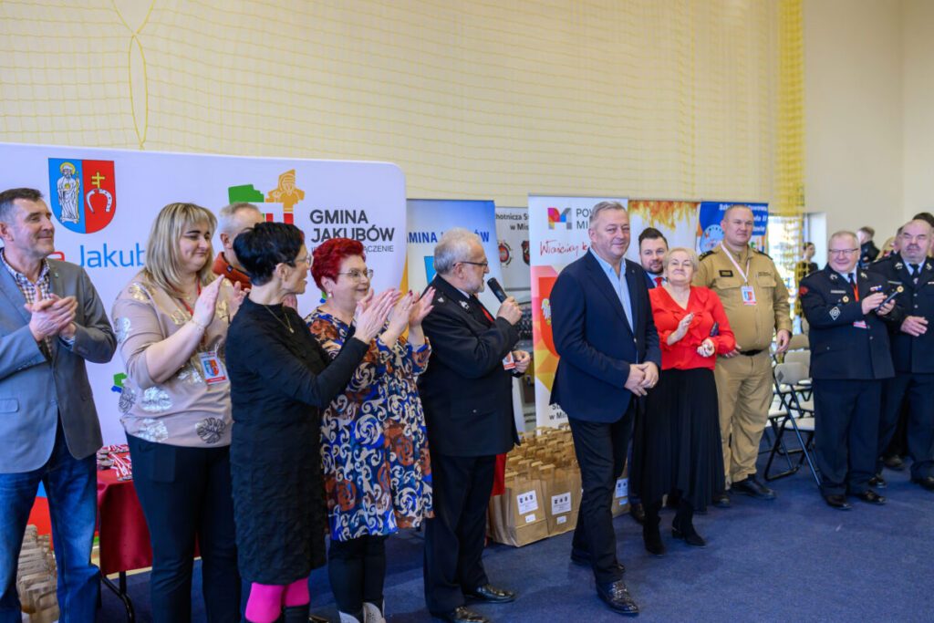 Grupa osób stoi przed tablicami z logami gminy na spotkaniu. Kolory: jasne tło, ubrania w ciemnych odcieniach. Część z nich klaszcze.