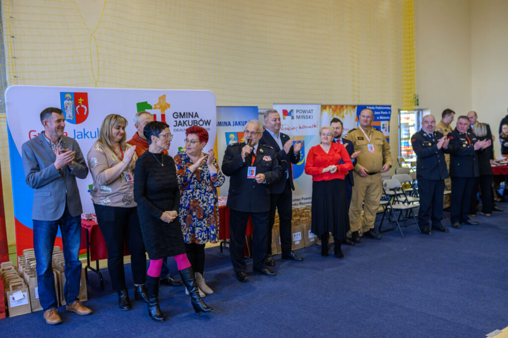 Grupa osób stoi przed banerami w jasnej sali. Dominują kolory: niebieski, czerwony, beżowy. Widoczne napisy Gmina Jakubów oraz Powiat Miński.
