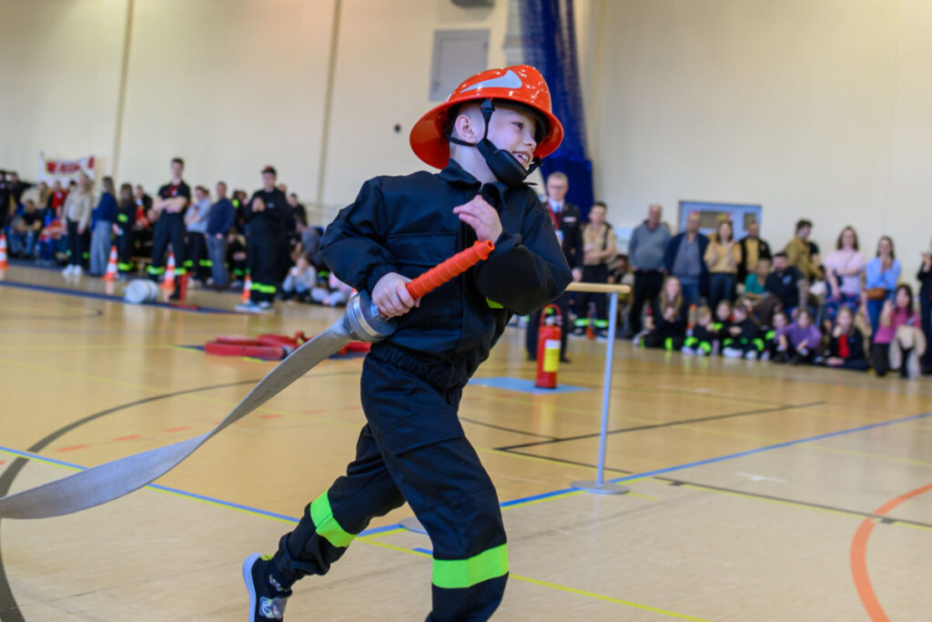 Chłopiec w strażackim stroju biega po sali gimnastycznej, trzymając wąż. Ma na sobie pomarańczowy kask. W tle widoczni są widzowie.