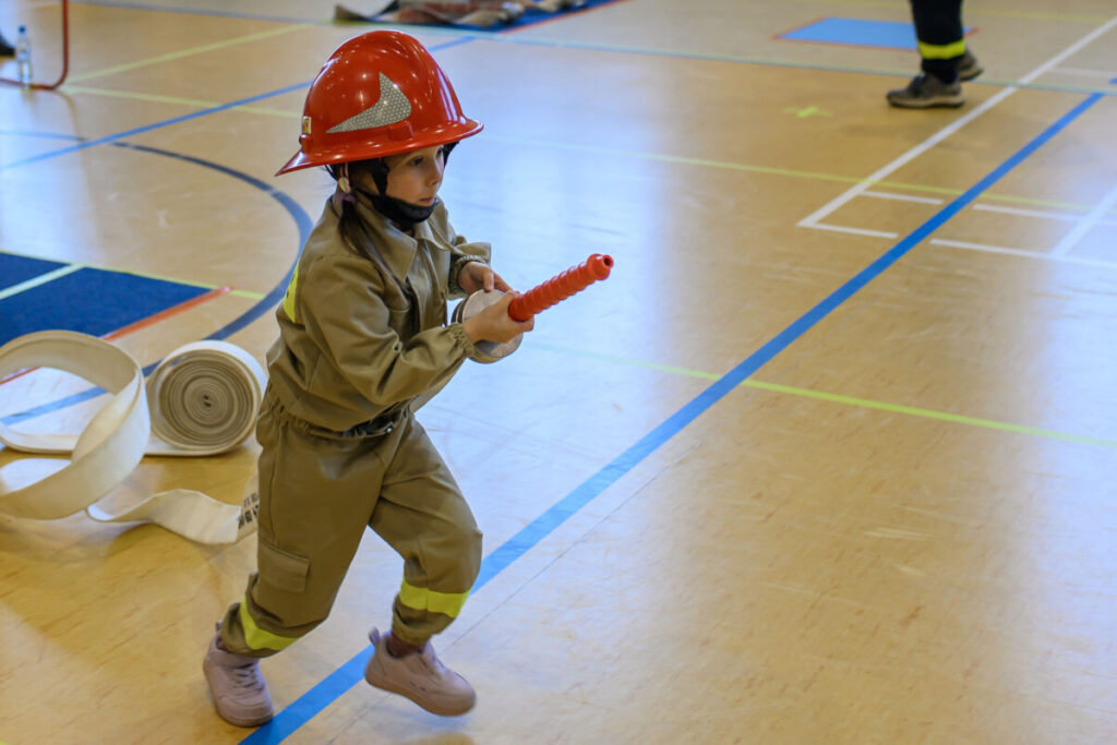 Dziecko w stroju strażaka z czerwonym kaskiem biega po sali gimnastycznej. W tle rozłożone węże. Podłoga jasna, sportowa.