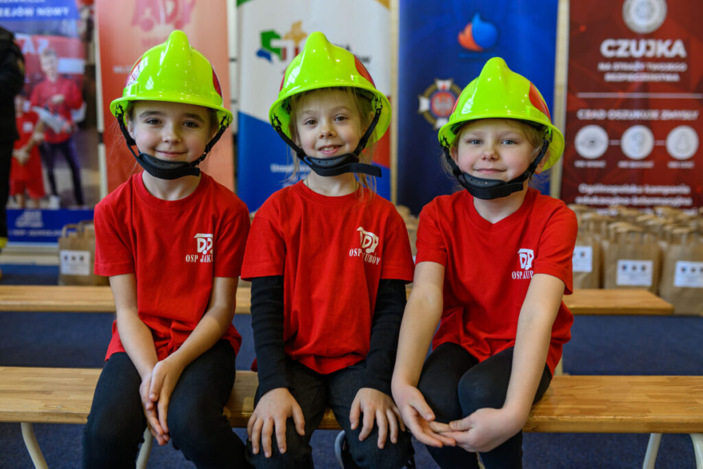 Trzy dzieci w zielonych kaskach i czerwonych koszulkach siedzą na ławce. W tle kolorowe banery i torby. Uśmiechają się.