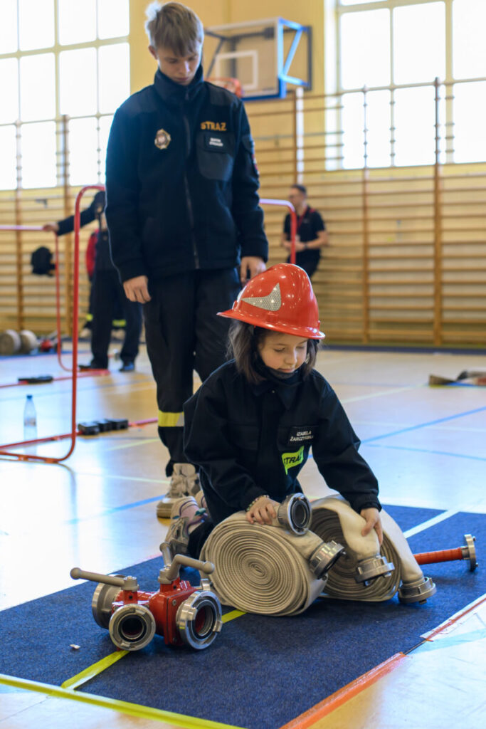 Młoda dziewczynka w czerwonym hełmie trzyma wąż strażacki. Obok niej stoi chłopiec w ciemnym mundurze. Tło to sala gimnastyczna z jasnymi ścianami.