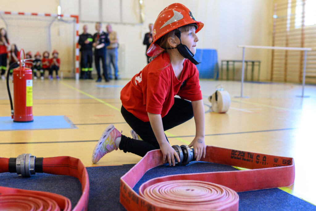 Małe dziecko w czerwonym kasku i koszulce trzyma węża strażackiego na hali sportowej. Tło jest jasne, z grupą ludzi w oddali. Na podłodze jasne panele, obok stoi gaśnica.