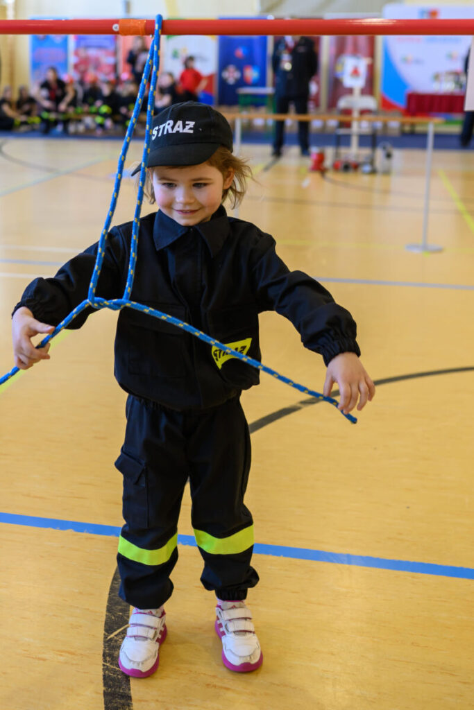 Dziewczynka w czarnym stroju strażaka trzyma linę na sali gimnastycznej. Tło to kolorowe banery i osoby zebrane w grupach. Podłoga jest jasna z niebieskimi liniami.