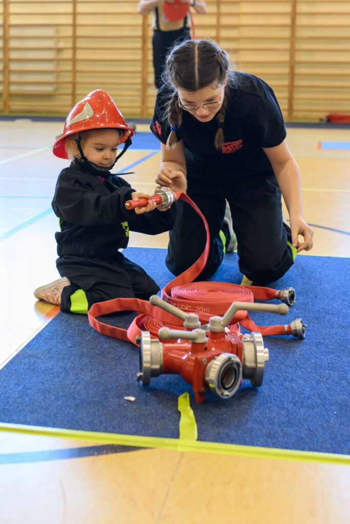 Dziecko w czerwonym hełmie strażackim i kobieta w czerni trzymają wąż strażacki na sali gimnastycznej. Kolorowy sprzęt leży na niebieskim dywanie. W tle są drewniane drabinki.