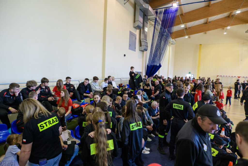 Na zdjęciu widać grupę ludzi w czarnych ubraniach z napisem STRAŻ, siedzących na niebieskich krzesłach. Tło stanowi jasna ściana sali z drewnianym sufitem i siatką sportową. Atmosfera jest gęsta, sugerująca