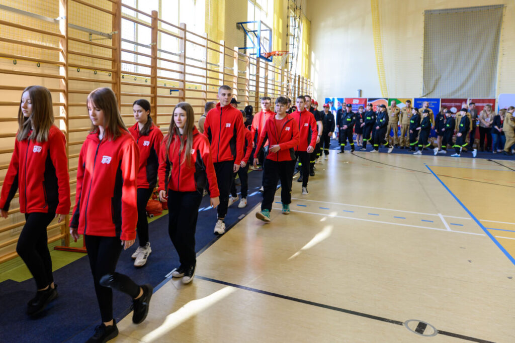 Grupa młodych ludzi w czerwonych kurtkach maszeruje po sali gimnastycznej. Podłoga jasna, ściany w tonacji beżowej. W tle stoją osoby w ciemnych mundurach.