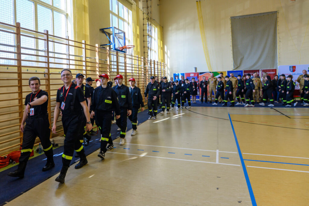 Grupa strażaków w ciemnych uniformach maszeruje w jasnej sali gimnastycznej. Po prawej tablice informacyjne. Drewniane drabinki na ścianie.