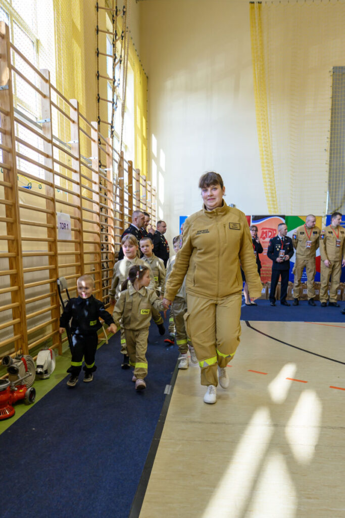 Grupa dzieci w strojach strażackich idzie po sali gimnastycznej. W tle drabinki i jasnożółte ściany. Obok dorosła osoba również w mundurze.