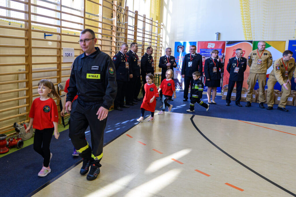 Dzieci i strażak w mundurze spacerują w sali gimnastycznej. W tle stoją dorośli w mundurach, obok są kolorowe banery. Podłoga jest jasna, na ścianie drewniane drabinki.