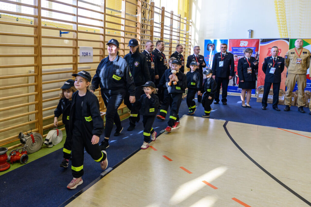 Na sali gimnastycznej grupa dzieci w strażackich strojach maszeruje, trzymając się za ręce. W tle stoją strażacy w mundurach. Jasne ściany i drewniane drabinki dominują w otoczeniu.