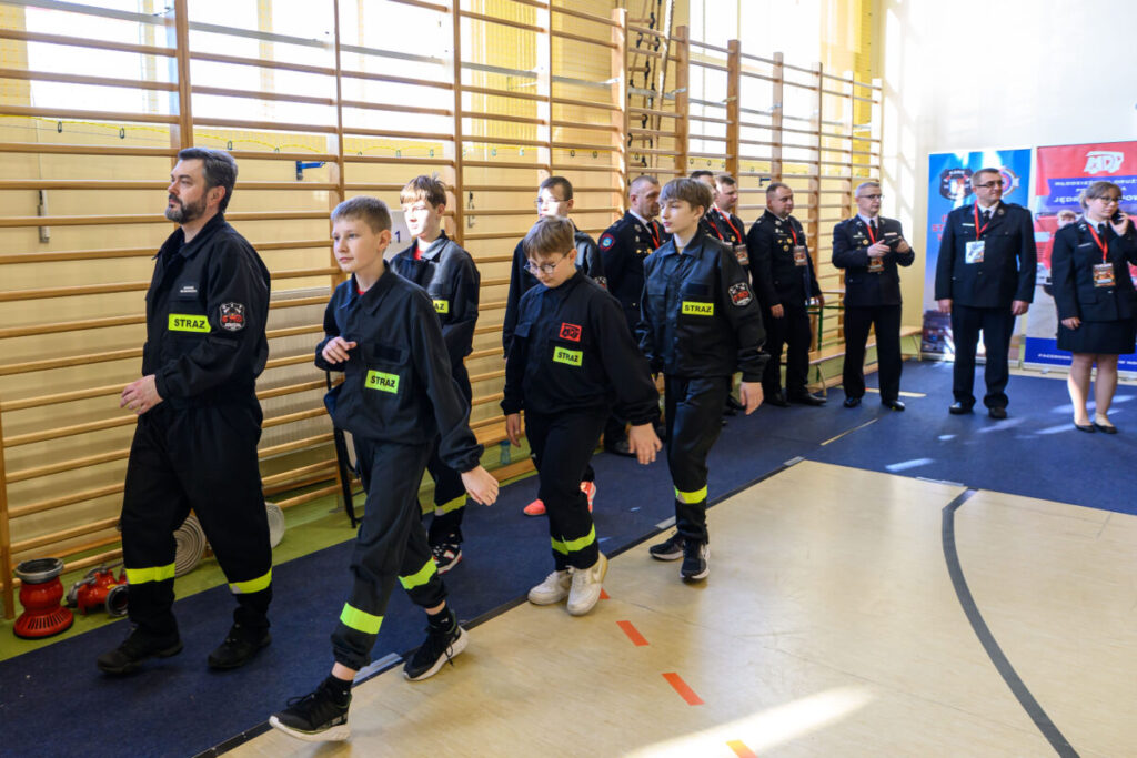 Grupa osób w czarnych kombinezonach strażackich maszeruje w sali gimnastycznej. Tło to drewniane drabinki i banery strażackie. Podłoże jest jasne, a na nim niebieski dywan.