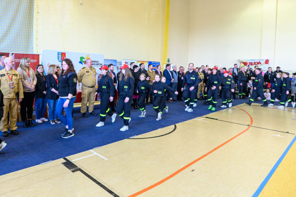 Grupa strażaków stoi i idzie po sali gimnastycznej w czarnych strojach. Tło jasne z żółtymi ścianami i banerami. Widać osoby w beżowych uniformach na niebieskim dywanie.