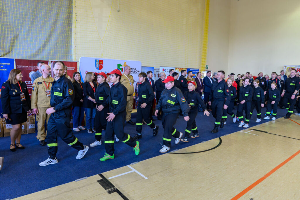 Grupa osób w strojach strażackich maszeruje w hali sportowej. Tło to ściany pokryte banerami i obserwujący widzowie. Kolory dominujące to czerń i czerwień.