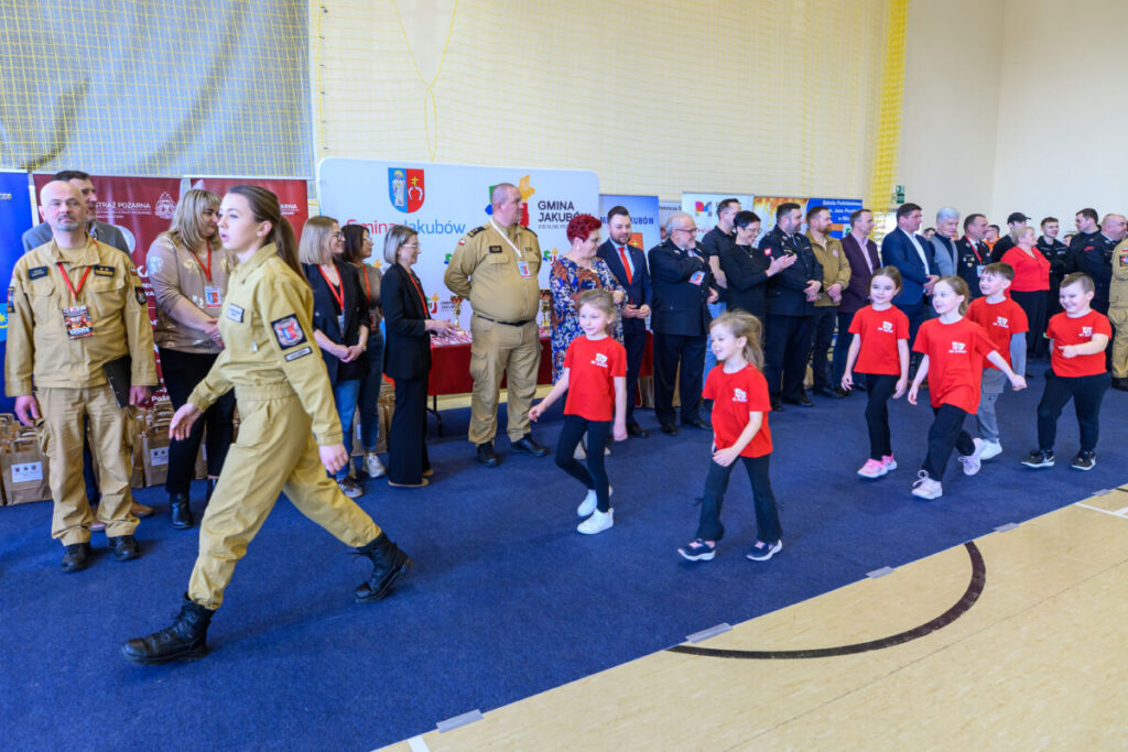 Grupa dorosłych i dzieci w sali; dzieci w czerwonych koszulkach maszerują; tło z banerami i żółtą ścianą.