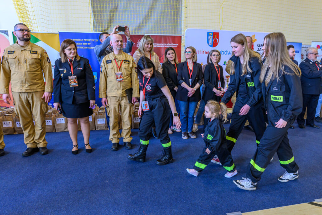 Grupa ludzi w mundurach strażackich i cywilnych stoi na niebieskim dywanie. W centrum jest kobieta z dzieckiem. Tło jest kolorowe z plakatami.