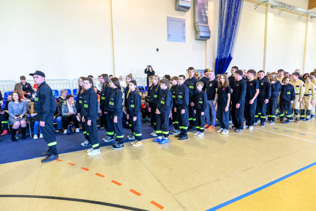 Grupa młodzieży w czarnych mundurach stoi w rzędach na sali gimnastycznej. Tło jasne, z niebieskimi krzesłami i widownią. Na podłodze linie wyznaczające strefy.