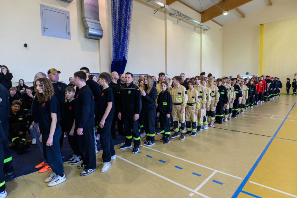 Grupa osób w sportowej sali ustawiona w rzędzie. Część ubrana jest w czarne stroje, inni w beżowe kombinezony. Tło to jasna ściana z elementami wentylacji.