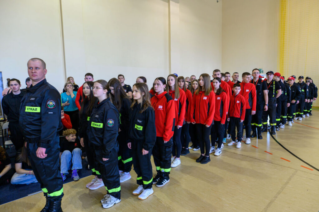 Grupa młodych ludzi stoi w rzędach w hali sportowej. Noszą czarne stroje strażackie i czerwone bluzy. Tło jest jasne, kremowe.