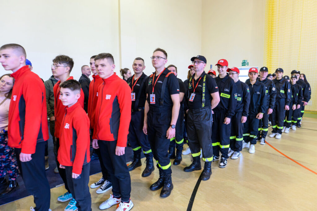 Na zdjęciu grupa młodych ludzi w czerwonych bluzach i strażacy w czarnych mundurach. Tło jest jasne, wewnątrz budynku. Kolory dominujące to czerwony, czarny i żółty.