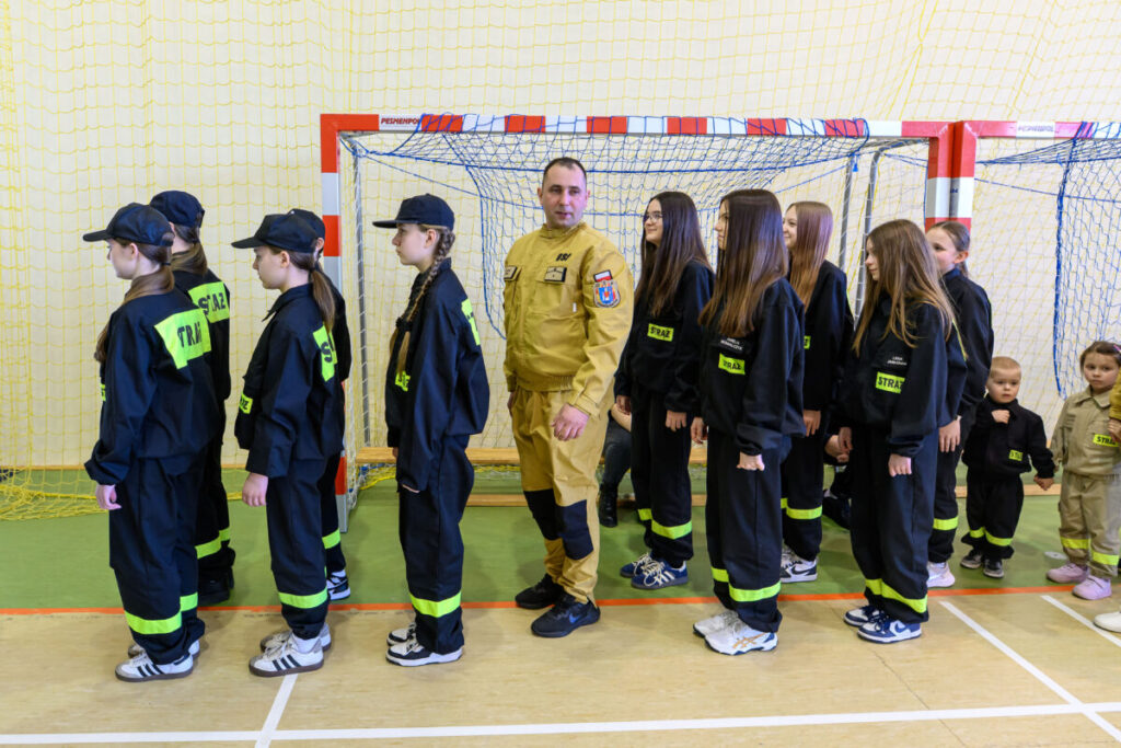 Na zdjęciu grupa dzieci w czarnych mundurach strażackich stoi w rzędzie na tle bramki do piłki ręcznej. Mundury mają jaskrawe, zielone naszywki z napisem STRAŻ. Tło to jasna ściana i siatka bramki z nie