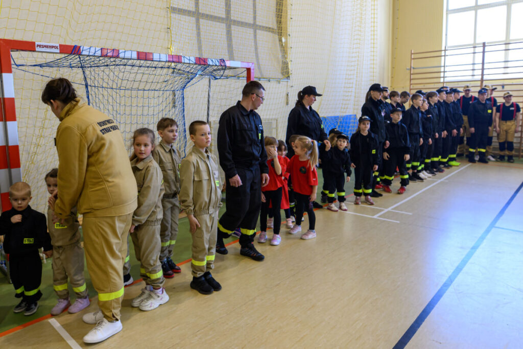 Grupa dzieci i dorosłych w strojach strażackich stoi w szeregu w sali gimnastycznej. Kolory strojów to beżowe i czarne. W tle widać bramkę i jasne ściany.