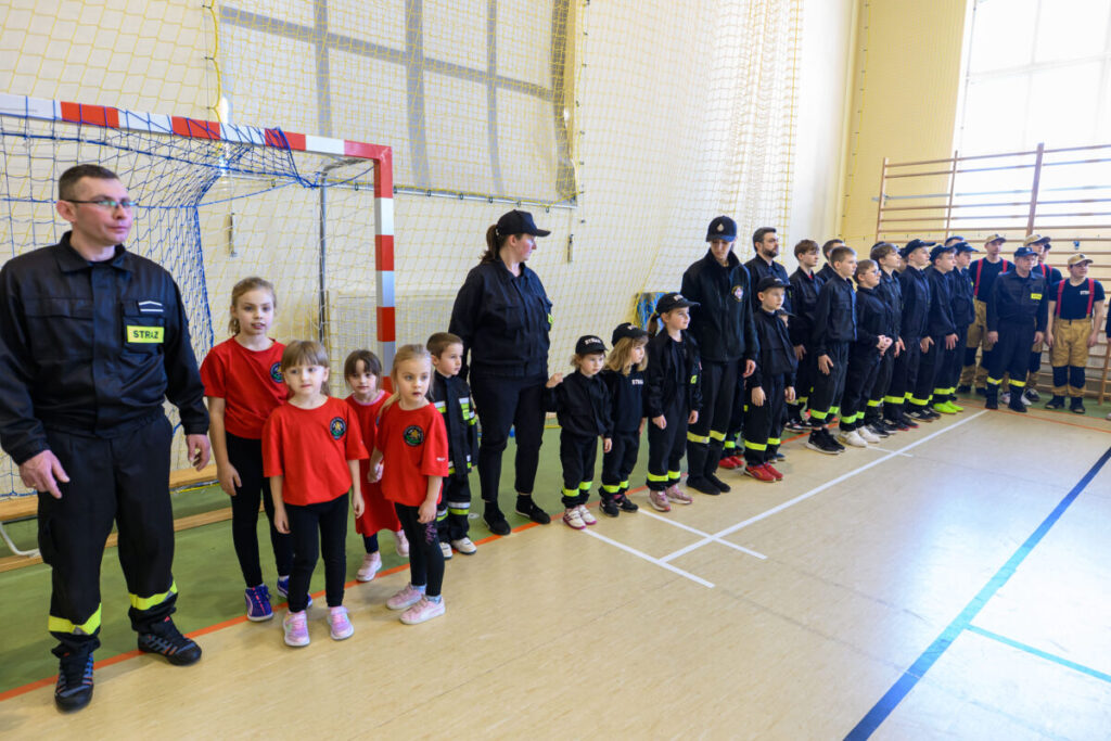 Dzieci i dorośli w strażackich strojach stoją w linii na sali gimnastycznej. Czerwone koszulki wyróżniają się na tle jasnych ścian i podłogi. W tle znajduje się bramka do piłki ręcznej i drewniane drabinki.