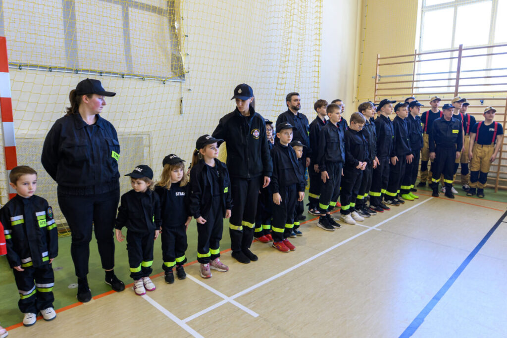 Grupa dzieci w mundurach strażackich stoi w linii w szkolnej sali gimnastycznej. Jednostajna kolorystyka czerni i zieleni. Za nimi widoczne są siatki i drabinki gimnastyczne.