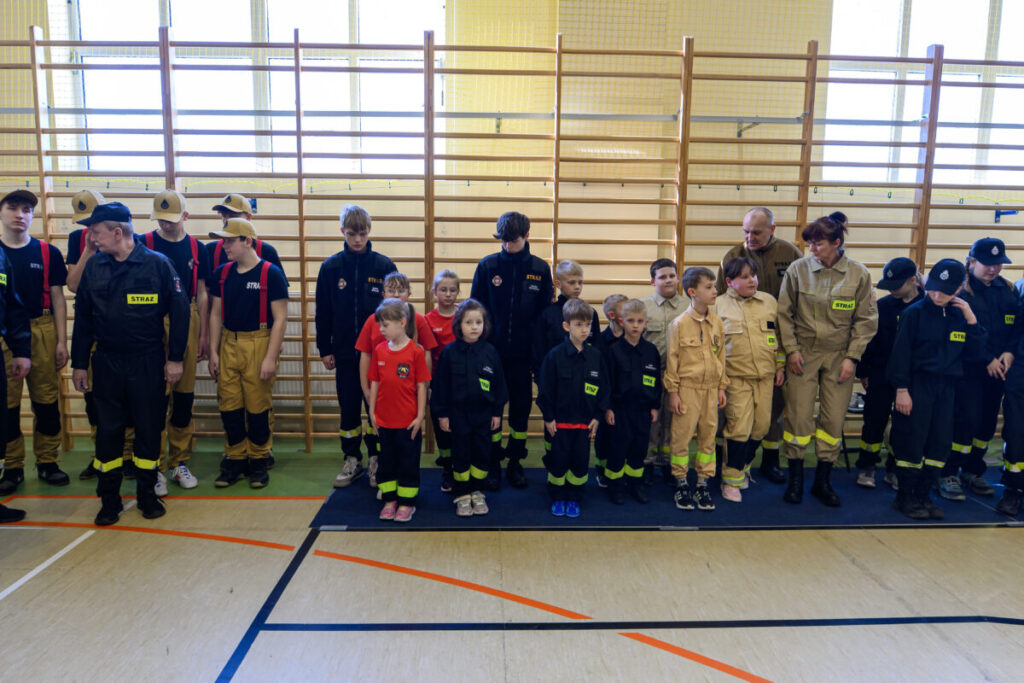 Grupa dzieci i dorosłych w mundurach strażackich stoi w sali gimnastycznej przed drabinką. Kolory: czarne i beżowe mundury. Tło: jasne ściany i podłoga z liniami sportowymi.