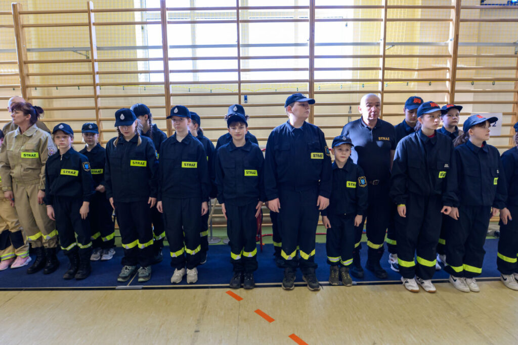 Grupa młodzieży w ciemnych uniformach strażackich stoi w rzędzie. Żółte napisy STRAŻ zdobią ich mundury. W tle jasne drabinki gimnastyczne.