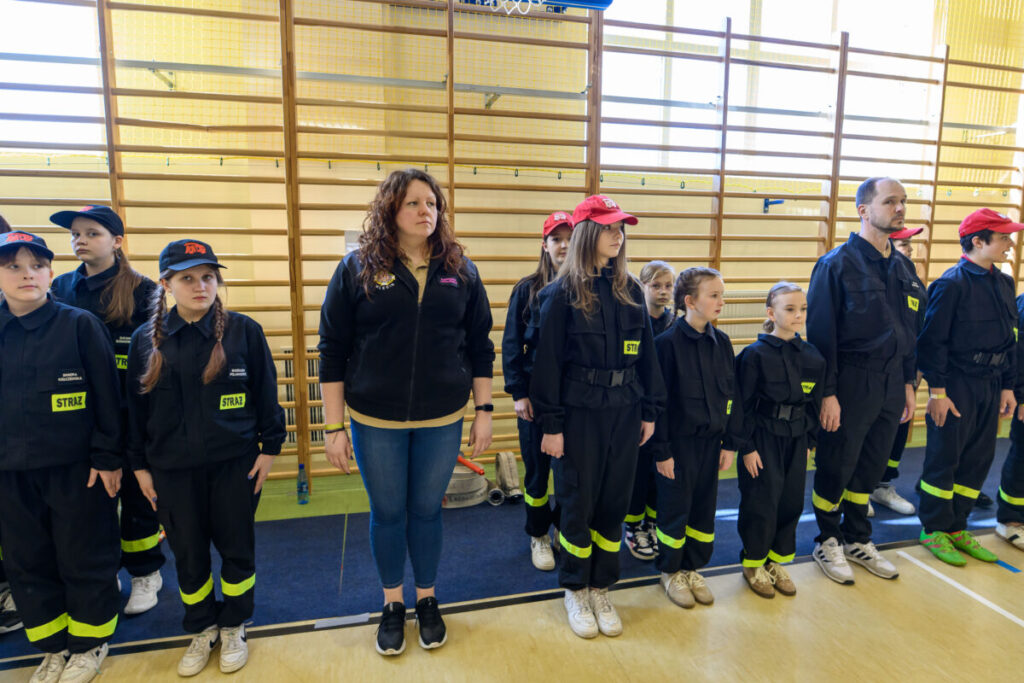 Grupa dzieci i dorośli w mundurach strażackich stoi w szeregu. Tło stanowią drabinki gimnastyczne w jasnej sali. Uczestnicy noszą czarne stroje z żółtymi naszywkami.