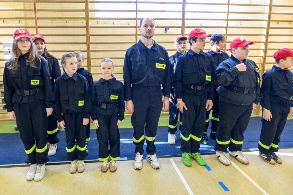 Grupa młodych strażaków i instruktor w ciemnych mundurach z żółtymi odznakami na sali gimnastycznej z jasnym drewnianym tłem. Dzieci mają czerwone czapki. Wszyscy stoją w równym szeregu.
