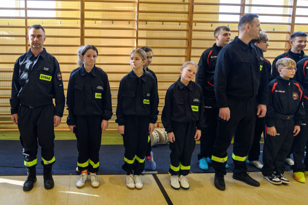 Grupa osób w mundurach strażackich stoi w sali gimnastycznej. Dominują kolory czarny i żółty. W tle widoczne są drewniane drabinki.