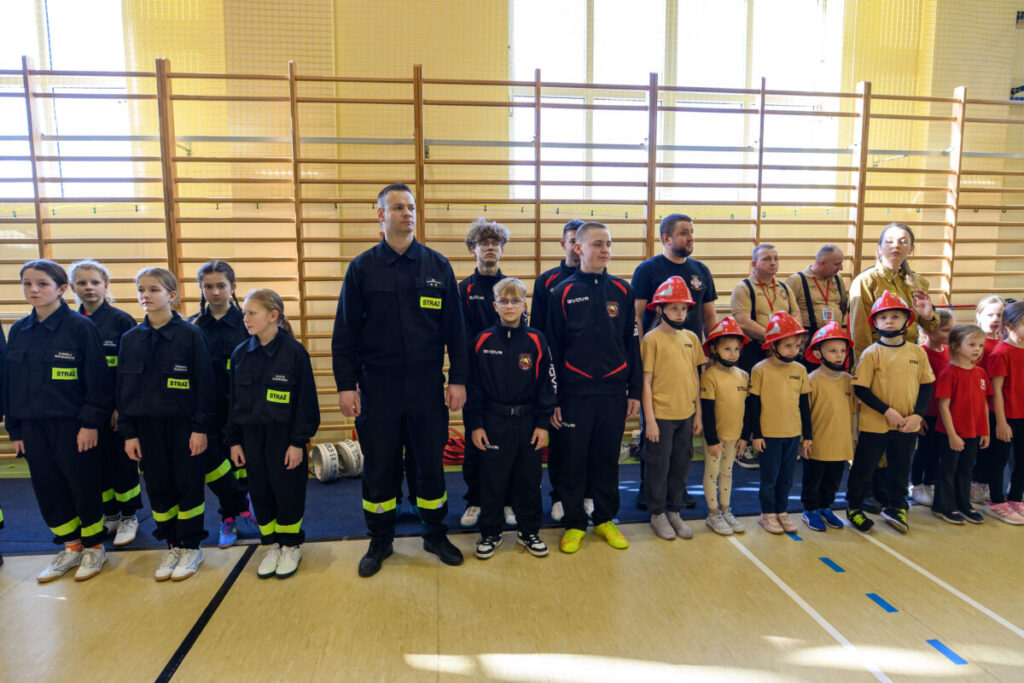Grupa dzieci i dorosłych w strojach strażackich stoi w szeregu na sali gimnastycznej. Niektóre dzieci noszą czerwone hełmy. W tle drabinki gimnastyczne i jasne ściany.