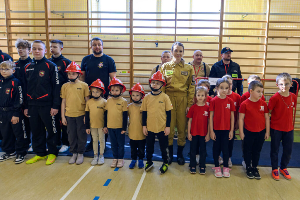 Grupa dzieci i dorosłych w strojach strażackich stoi w sali gimnastycznej. Dzieci mają czerwone hełmy, a dorośli ciemne uniformy. Tło stanowi drewniana drabinka ścienna.