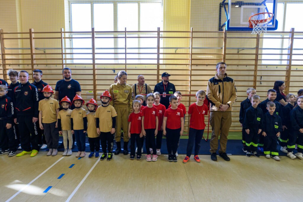 Grupa dzieci i dorosłych w strojach strażackich stoi w sali gimnastycznej. Tło stanowią drabinki gimnastyczne i kosz do koszykówki. Przeważają kolory czerwony i beżowy.