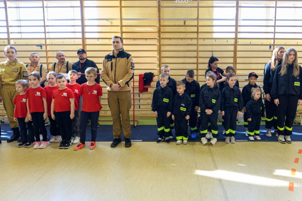 Na obrazie grupa dzieci i dorosłych pozuje w sali gimnastycznej. Dzieci noszą czerwone koszulki lub czarne stroje strażackie. W tle znajdują się drewniane drabinki na ścianie.