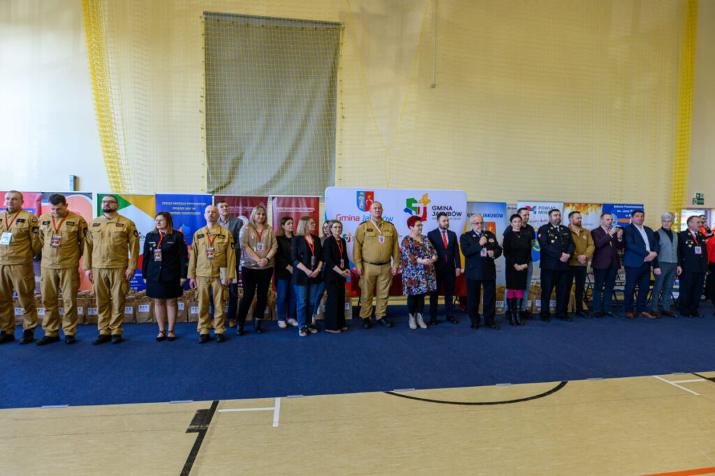 Grupa ludzi stoi w szeregu na hali sportowej, część w mundurach. W tle widoczne są kolorowe banery. Podłoga jest drewniana, a ściany jasne.