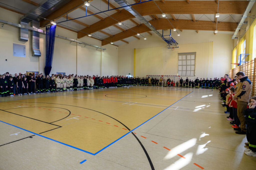 Ludzie w strojach strażackich stoją w rzędach na hali sportowej. Podłoga beżowa z liniami do gier, ściany jasne. Sufit z drewnianymi belkami.
